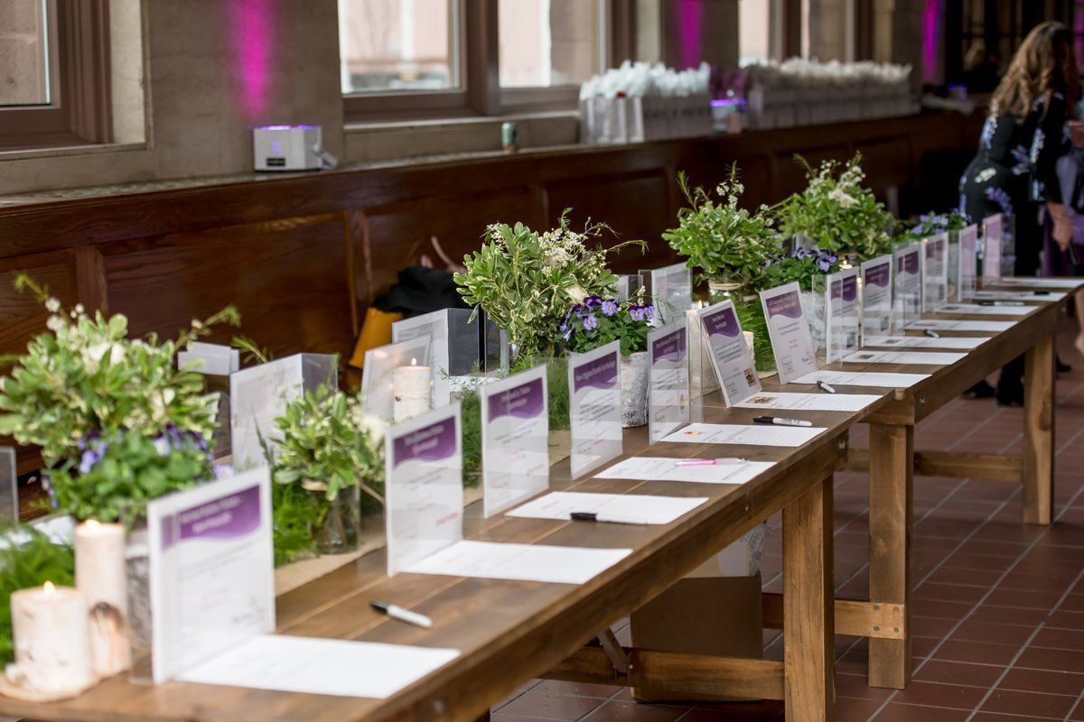 Long wooden table set with white cards, purple signs, and small flower arrangements in a hall