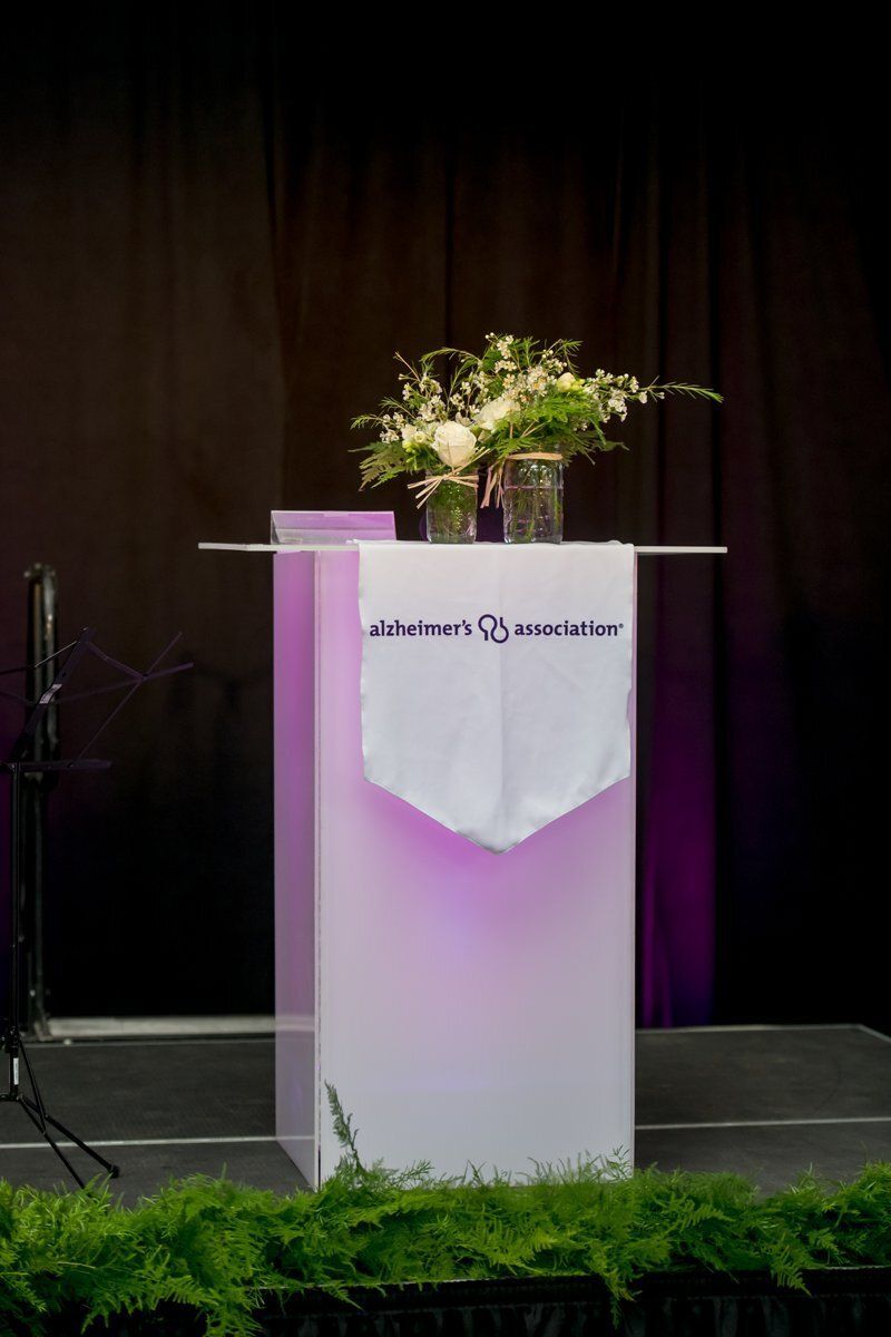 Flower arrangement on a lit white podium on a stage with black curtains and greenery in front