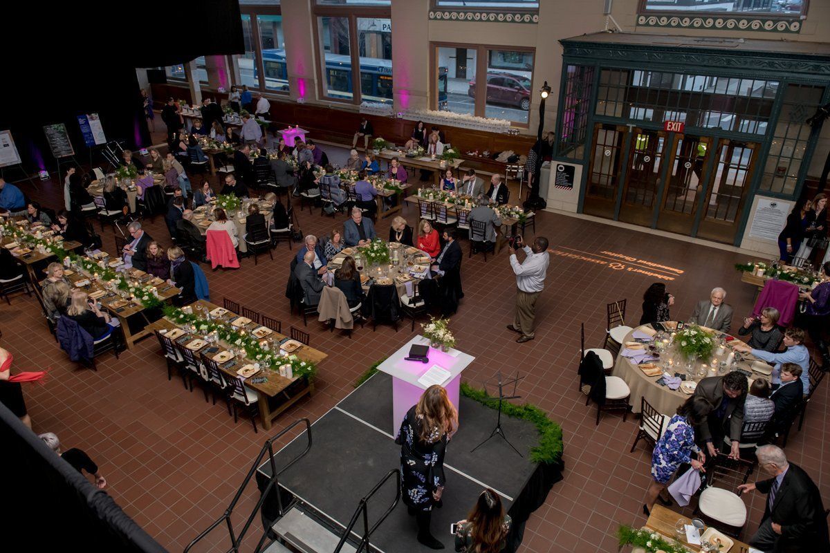 Crowded indoor banquet in a brick atrium, with round tables, a stage, and guests seated for a meal