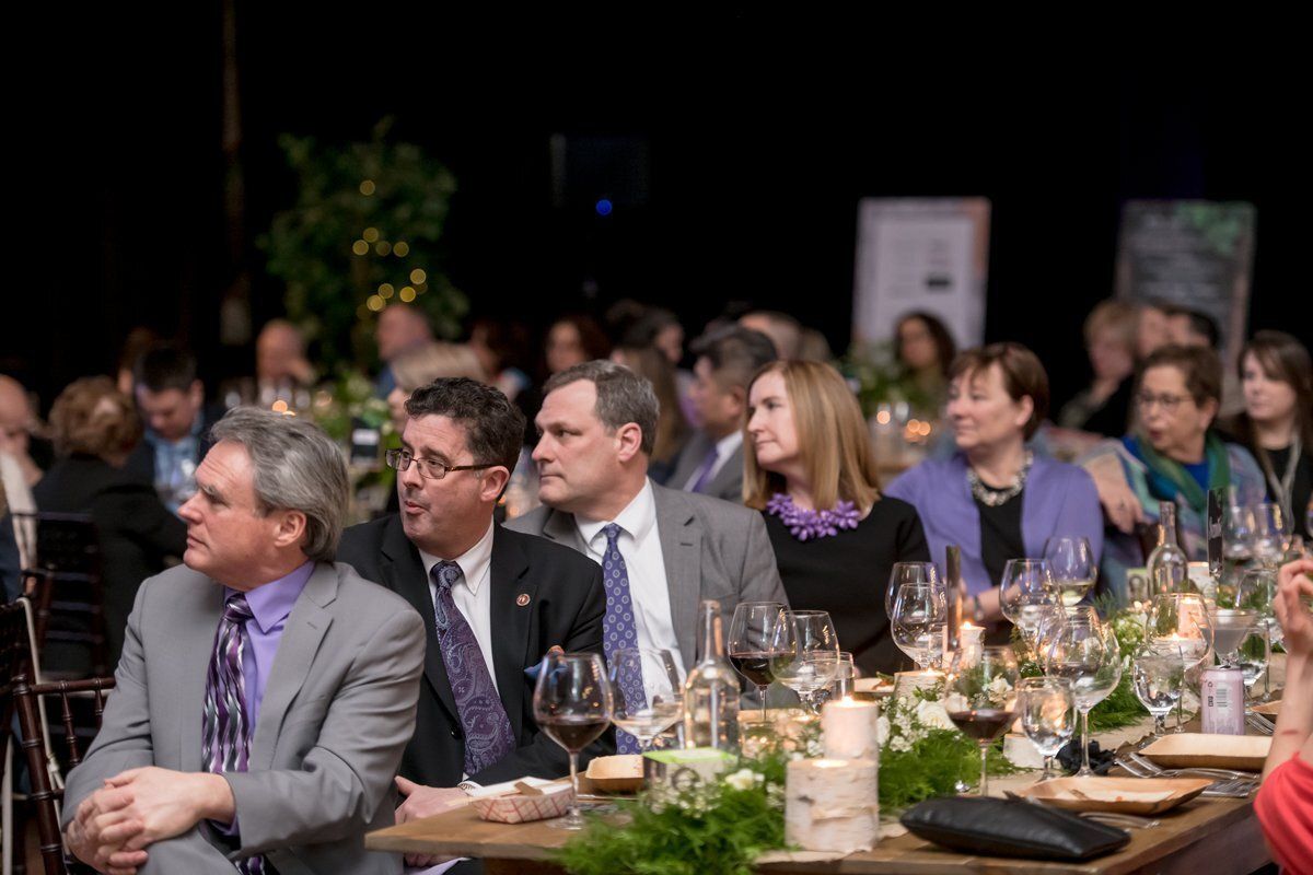 Guests seated at a banquet table, listening attentively in a dimly lit event hall