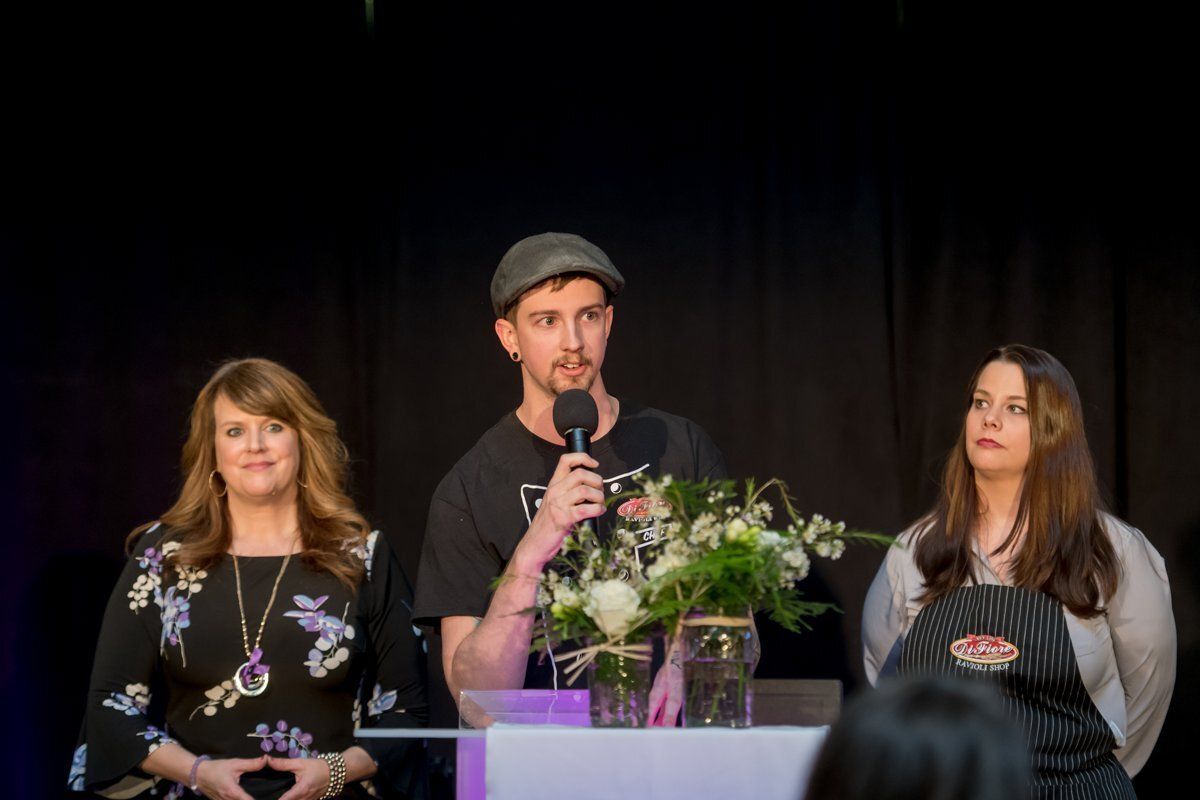 Three people stand at a podium on a stage, with a man speaking into a microphone.