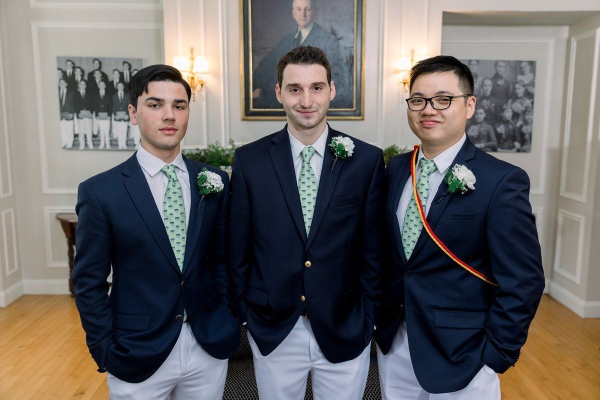 Three men in navy blazers and light pants posing indoors at a formal event