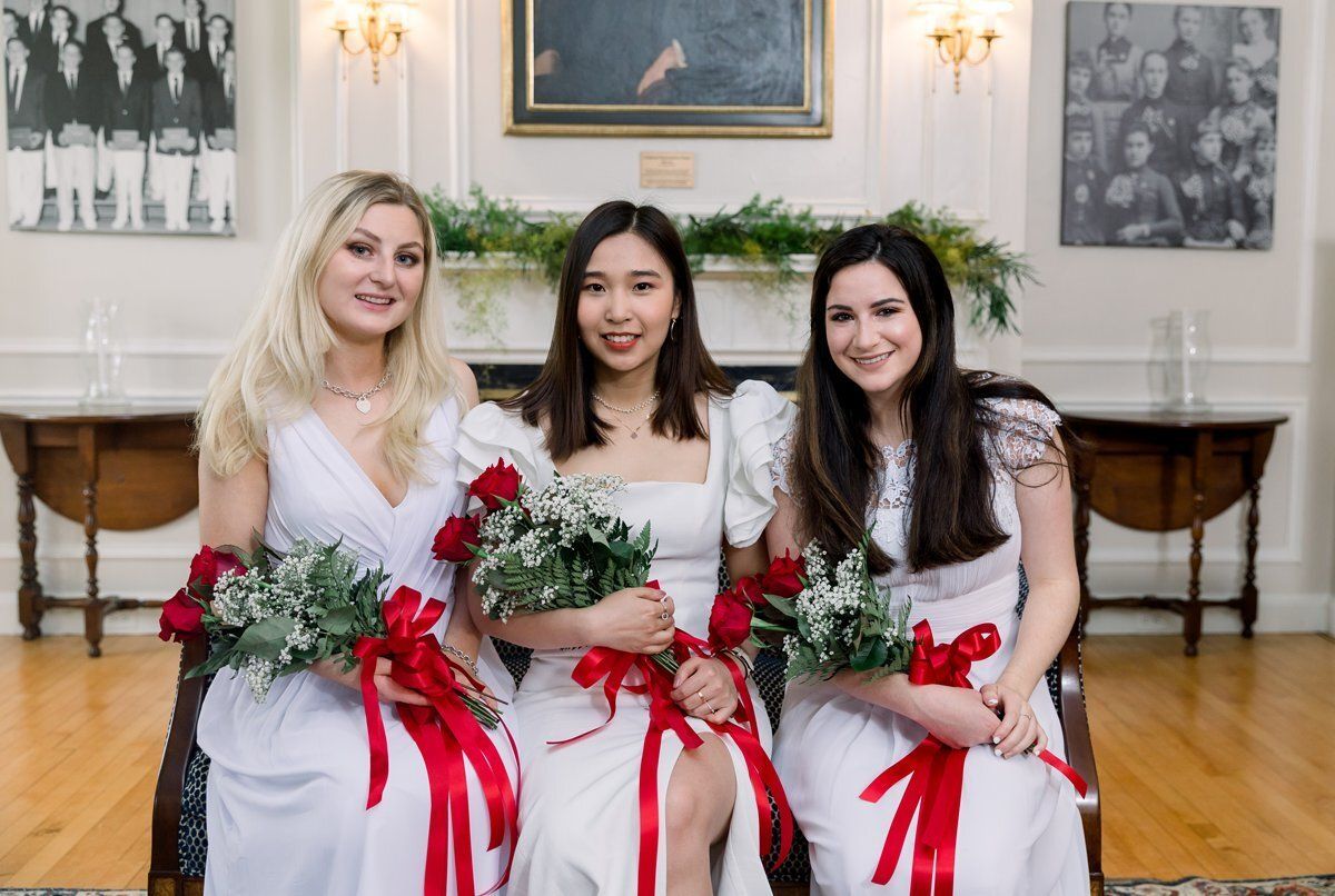 Three women in white dresses with red ribbons sit holding flower bouquets in a formal room.