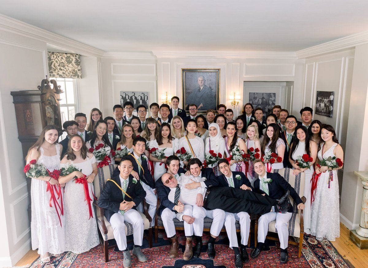 Large group posing in formal attire in a bright living room, some holding bouquets, with seated and standing rows