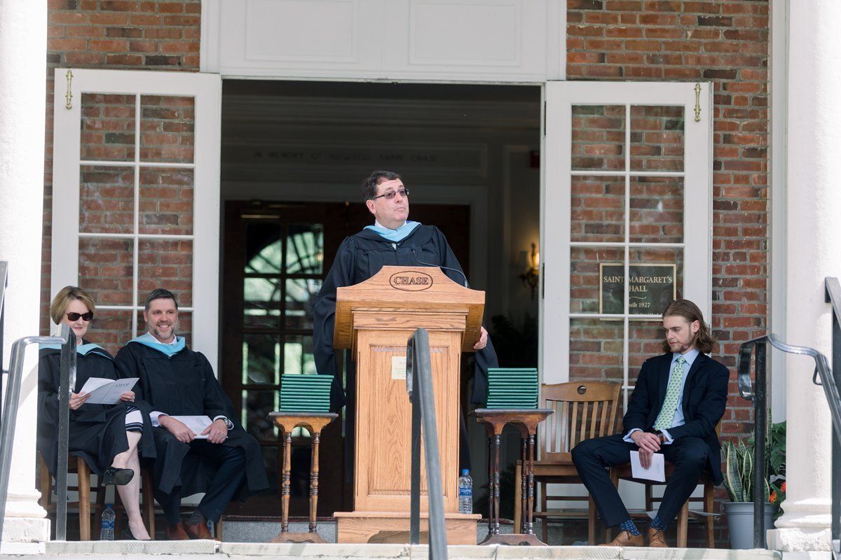 Speaker at podium addressing seated audience outside a brick building