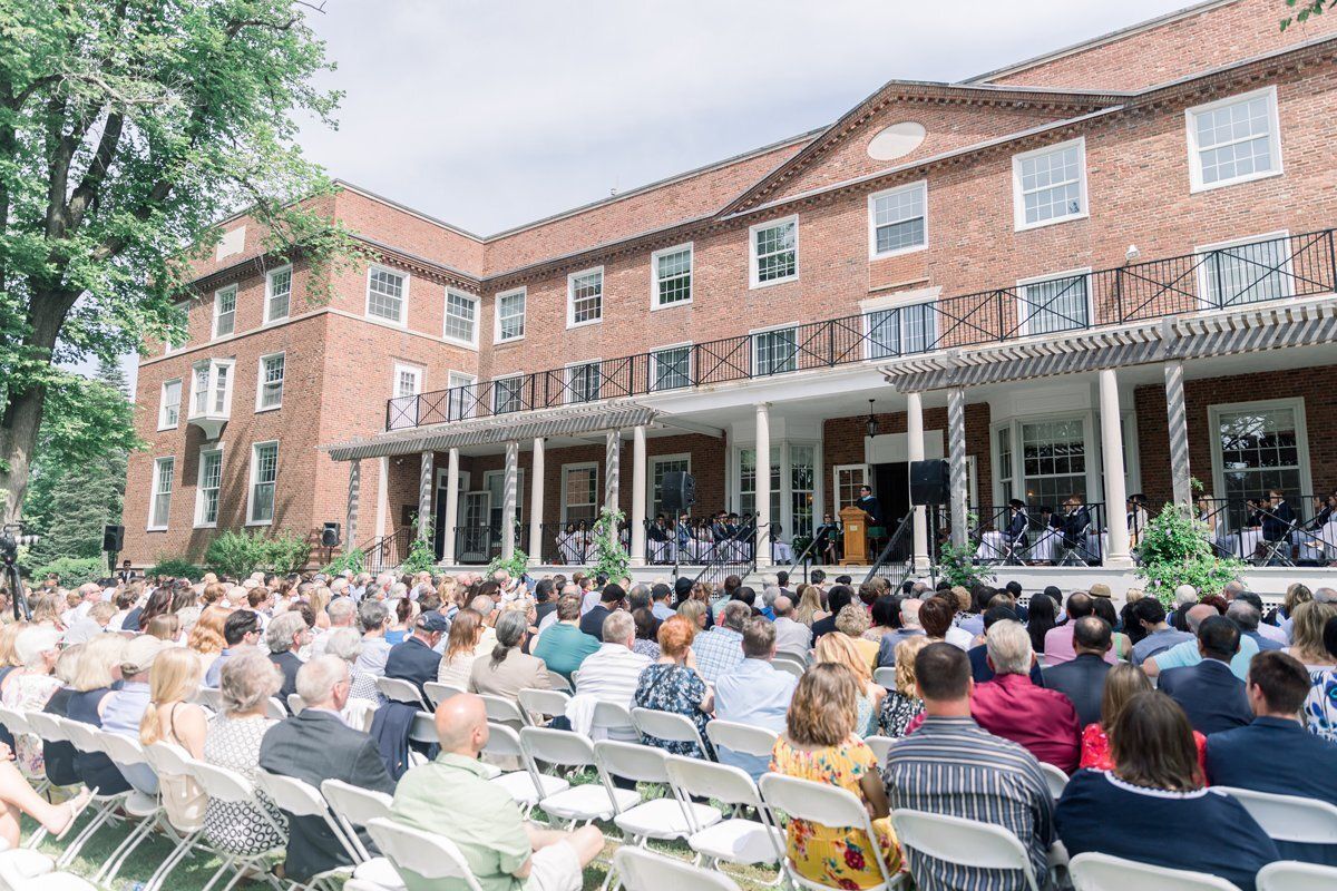 Large crowd seated outdoors at a brick building, watching a gathering on the patio