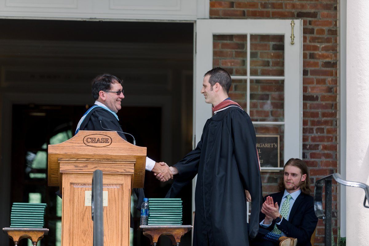 Two men shake hands at a graduation podium outside a brick building while another man claps nearby.