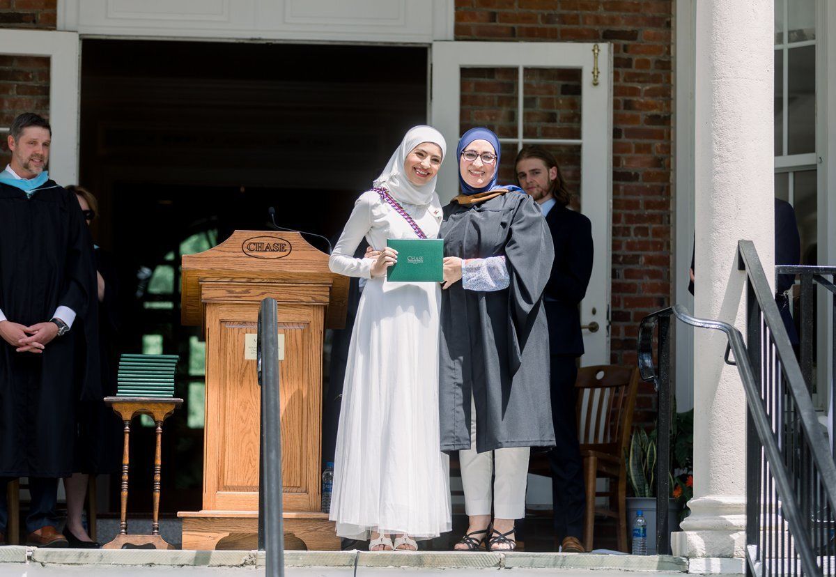 Two women in graduation attire pose on a building steps, holding a certificate folder.
