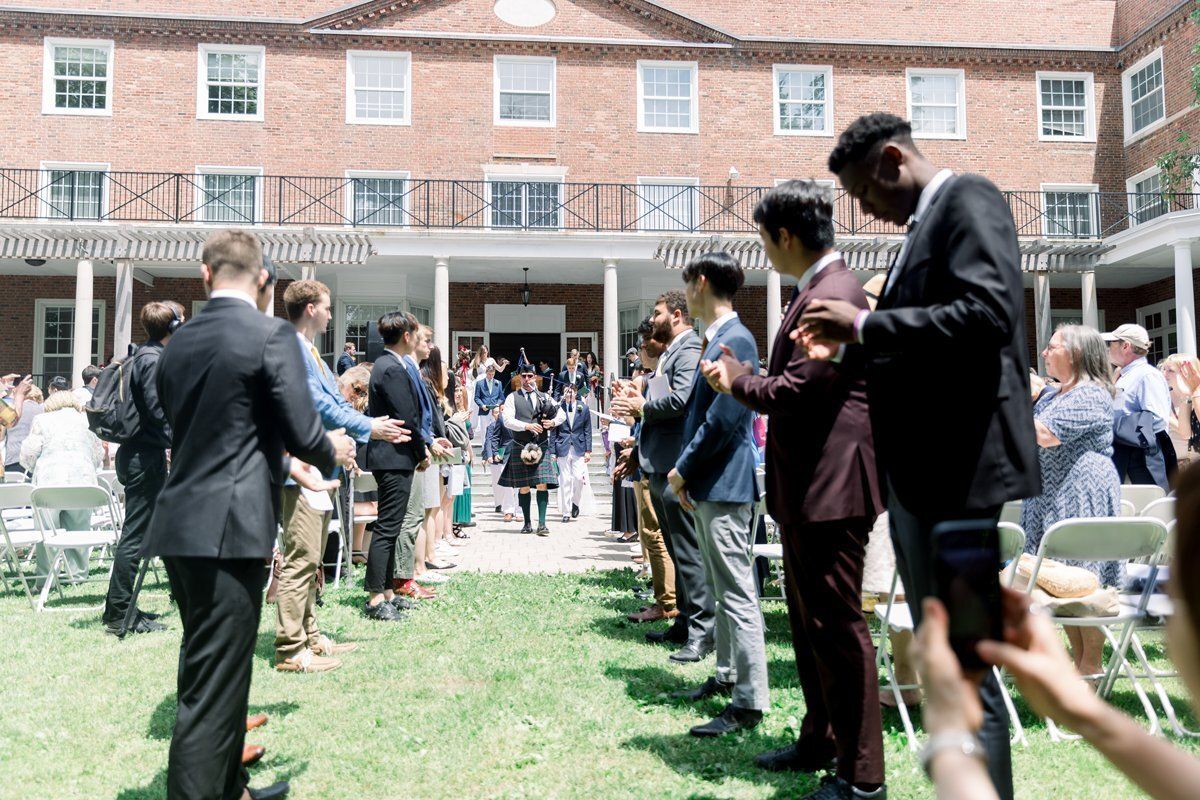 Guests in suits stand on a lawn at an outdoor wedding ceremony in front of a brick building.