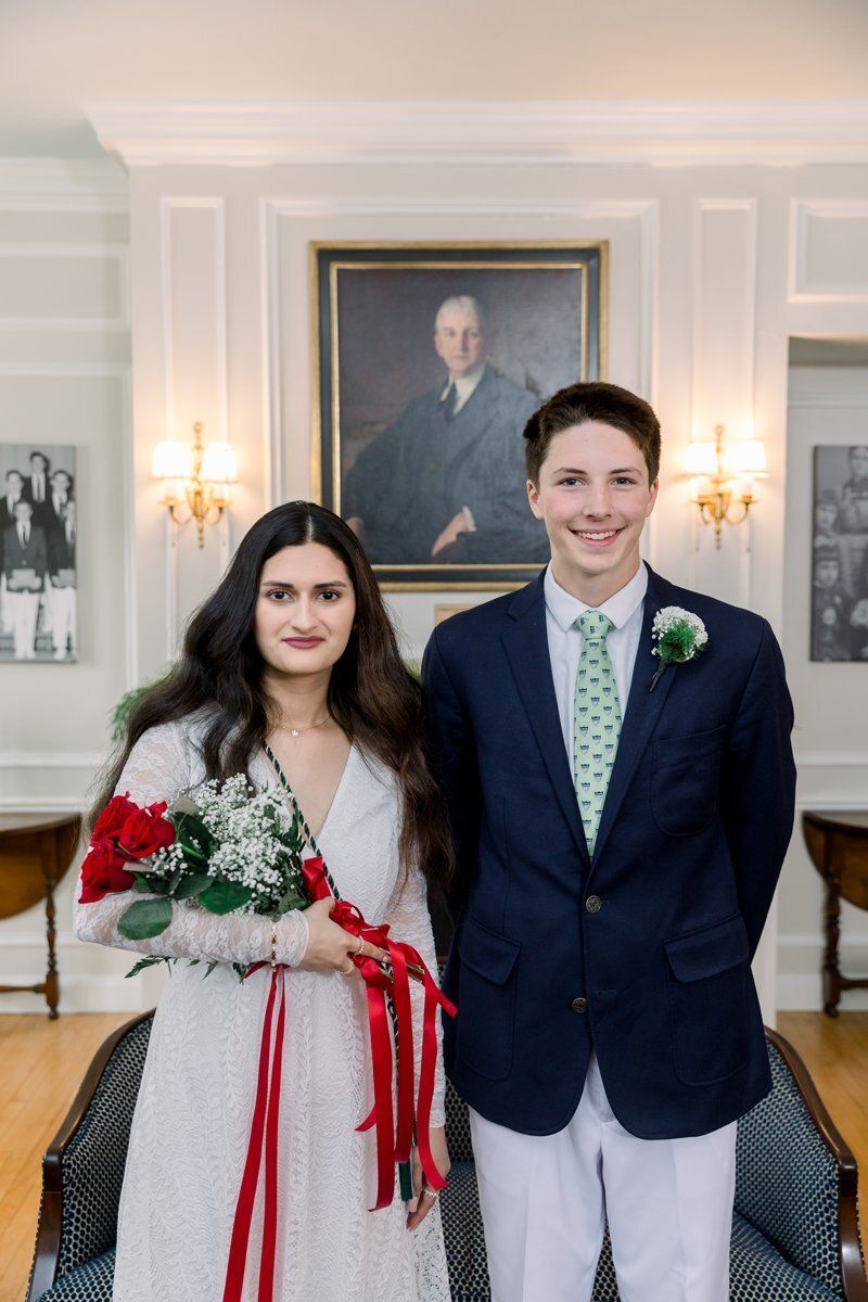 Couple posing indoors at a formal event, holding red and white flowers before a portrait wall