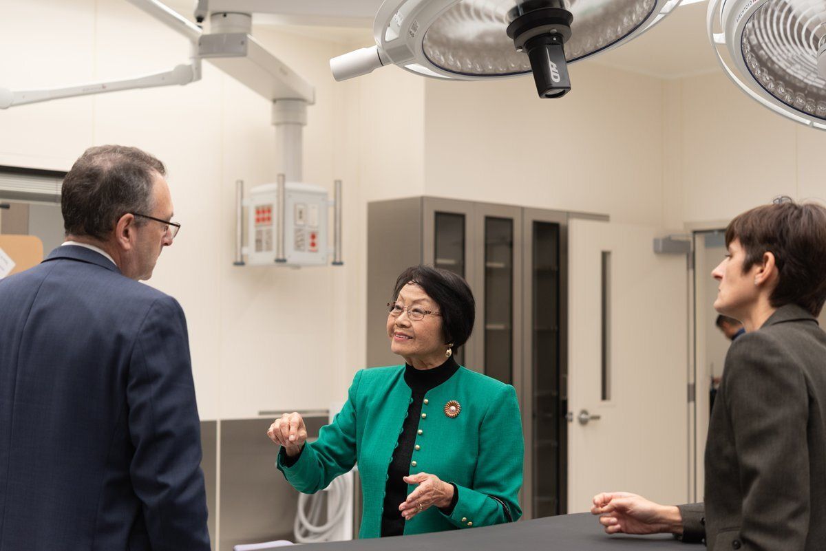 Three people talking in a medical room, with surgical lights overhead and a woman gesturing in green.