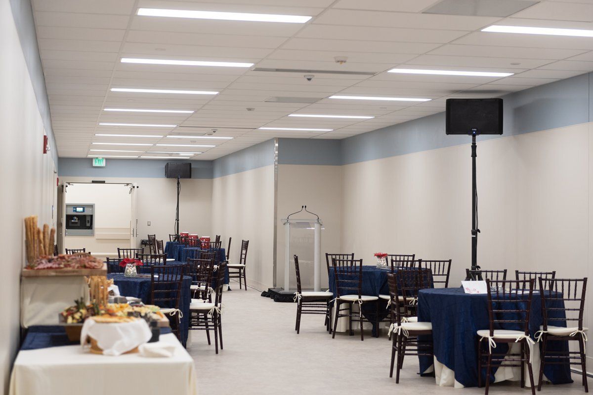 Large banquet hall with round tables, black chairs, blue tablecloths, and fluorescent ceiling lights