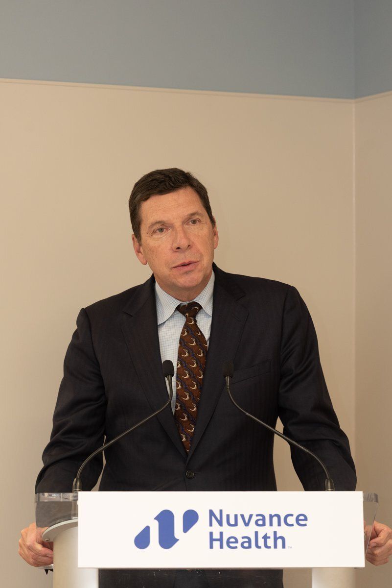 Man in dark suit speaking at a podium with Nuance Health logo against a plain wall.