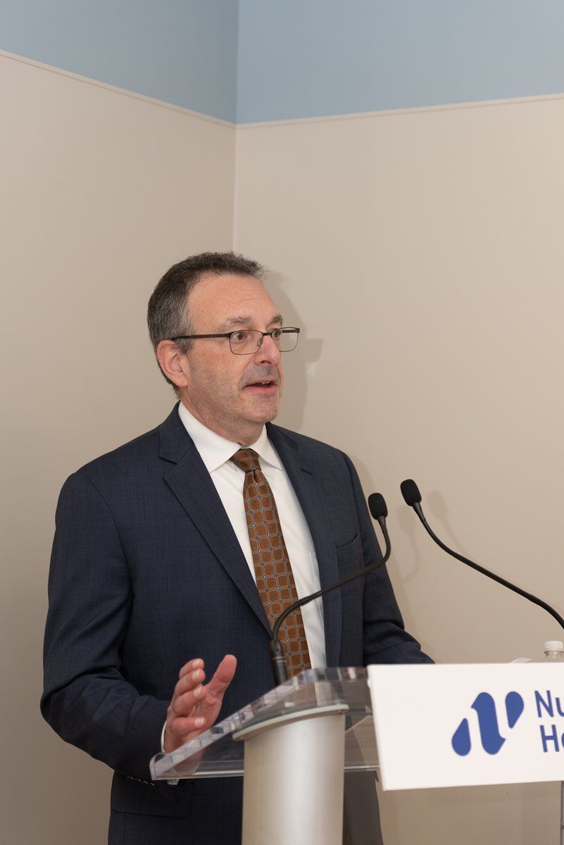 Man speaking at a podium with microphones in a beige room, wearing a navy suit and tie