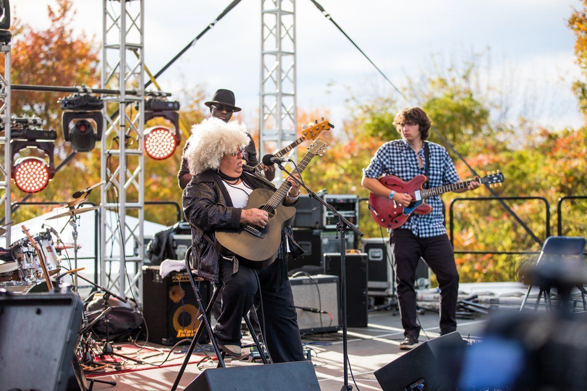 Musicians performing outdoors on stage, playing guitars and drums under bright daylight.