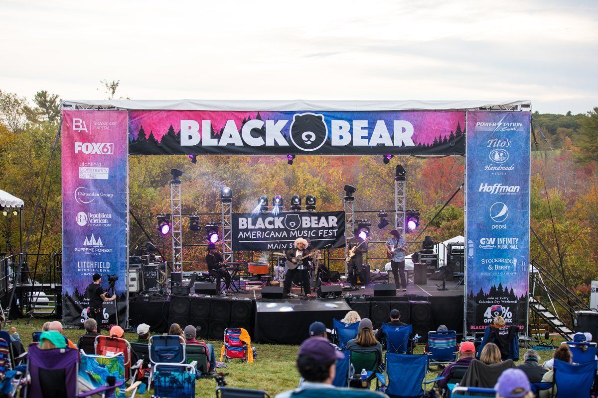 Outdoor concert stage with BLACK BEAR banner and seated audience at sunset