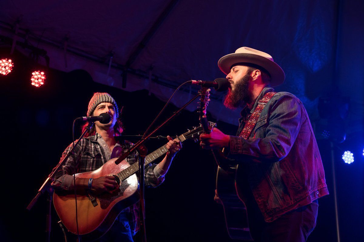 Two musicians perform onstage under purple and blue lights, one singing into a microphone, one playing guitar.