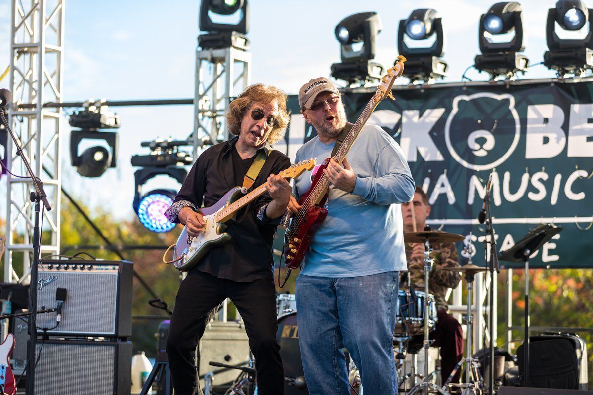 Two musicians playing guitars on an outdoor stage with lights and a music festival backdrop