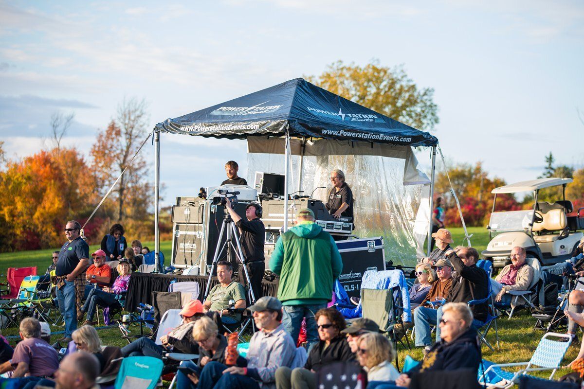 Crowd seated outdoors watching a live performance on a small stage under a blue canopy