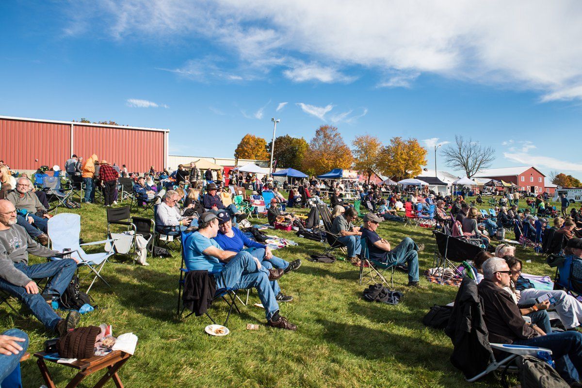 Crowd seated on folding chairs in a grassy field at an outdoor event under a blue sky