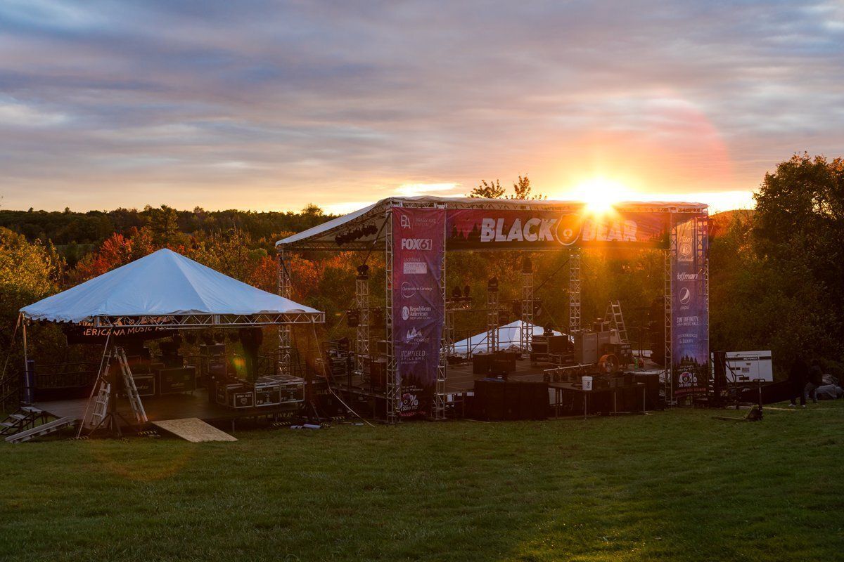 Outdoor stage with a black backdrop at sunset beside a white tent on a grassy field