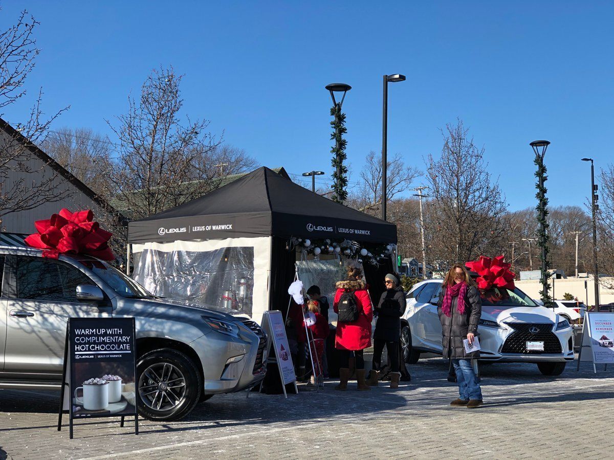 People gathered at an outdoor tent event with cars decorated with large red bows.