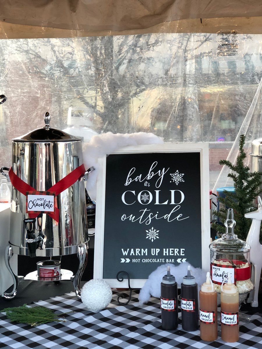 Hot cocoa bar with silver dispenser, mugs, chalkboard sign, and syrups on a plaid tablecloth.