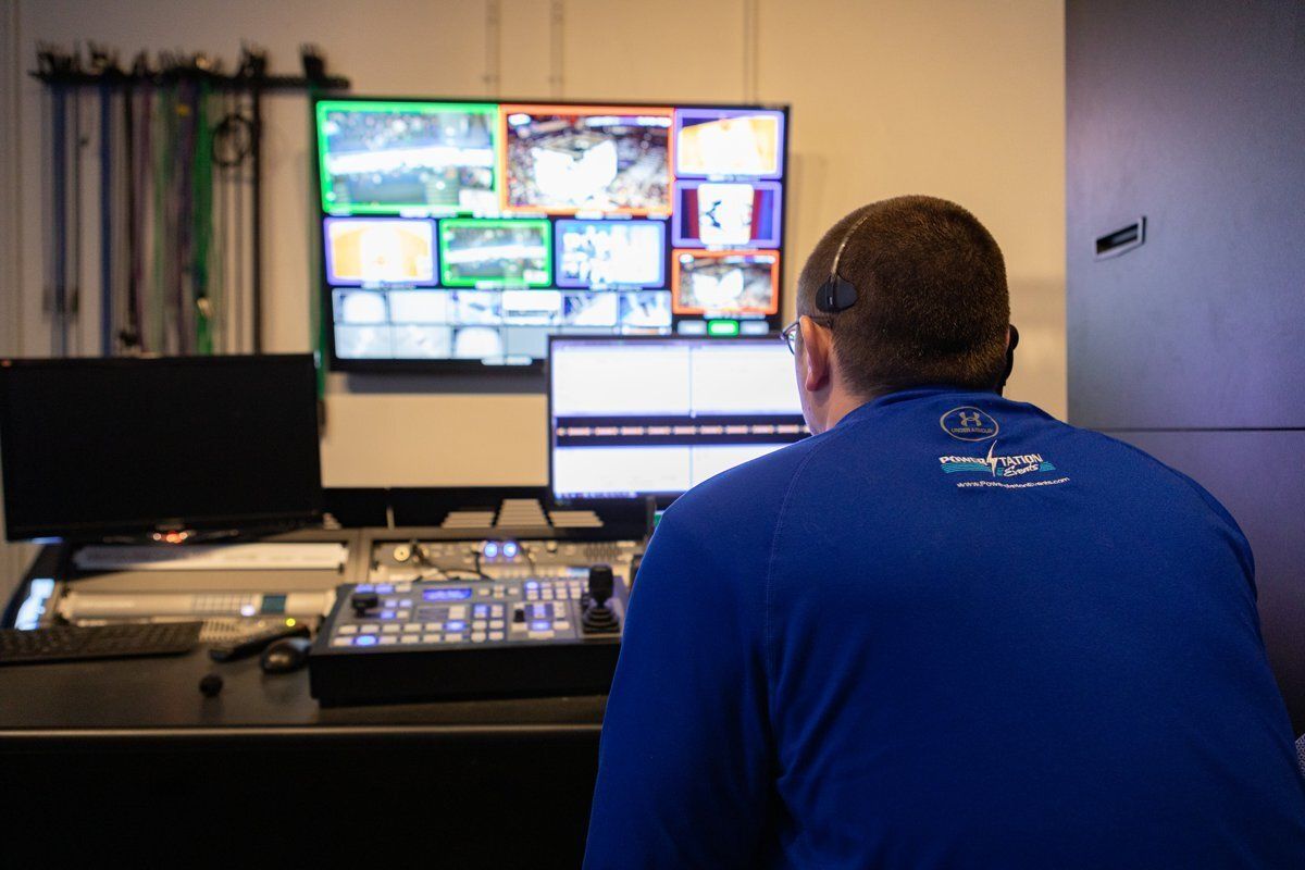Person at a control desk watching multiple monitors in a dimly lit operations room.