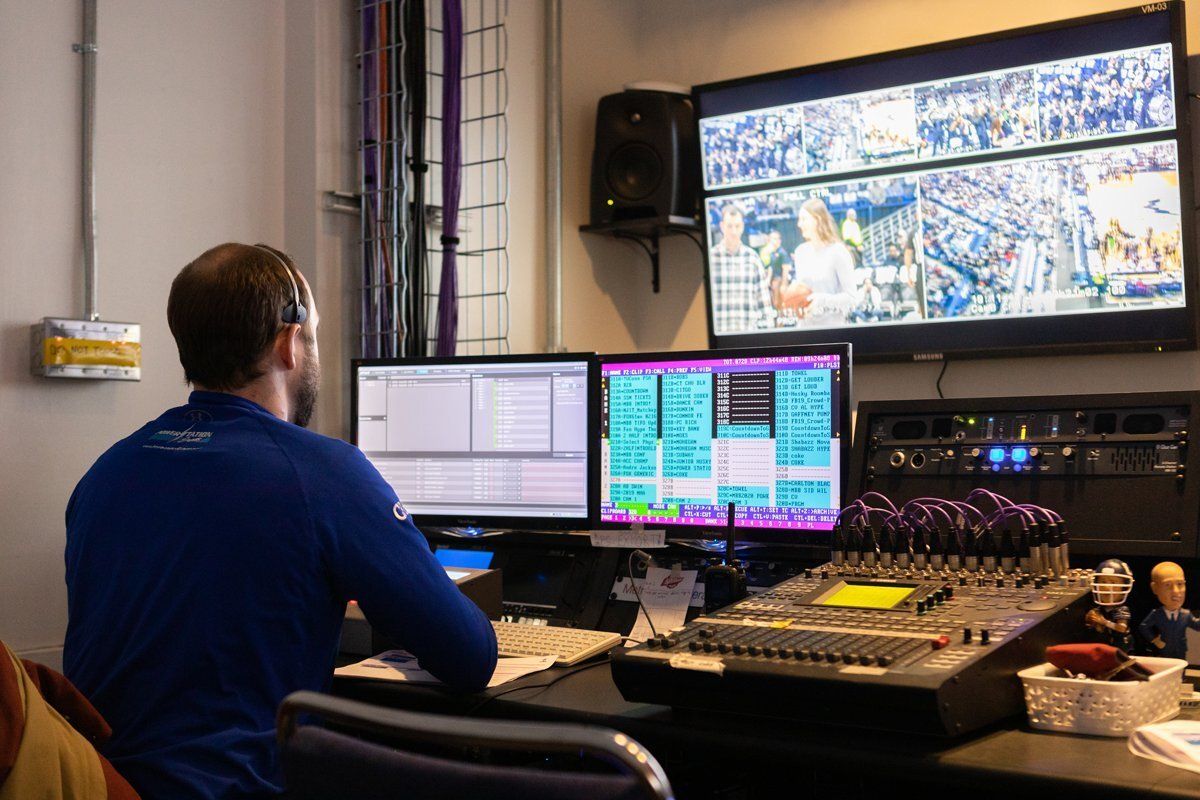 Person at an audio/video control desk with multiple monitors and a large wall display showing a live broadcast