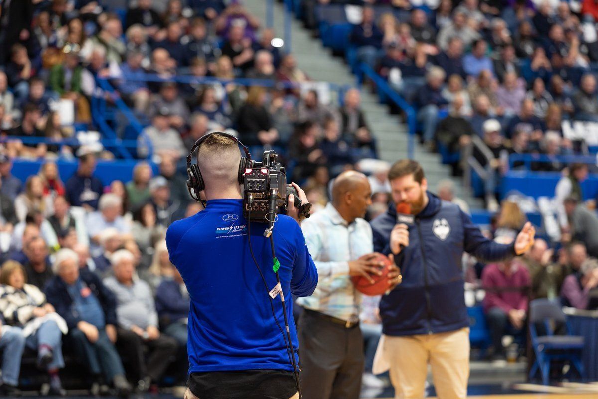 Camera operator films basketball players on court during a game, with crowd in the stands.