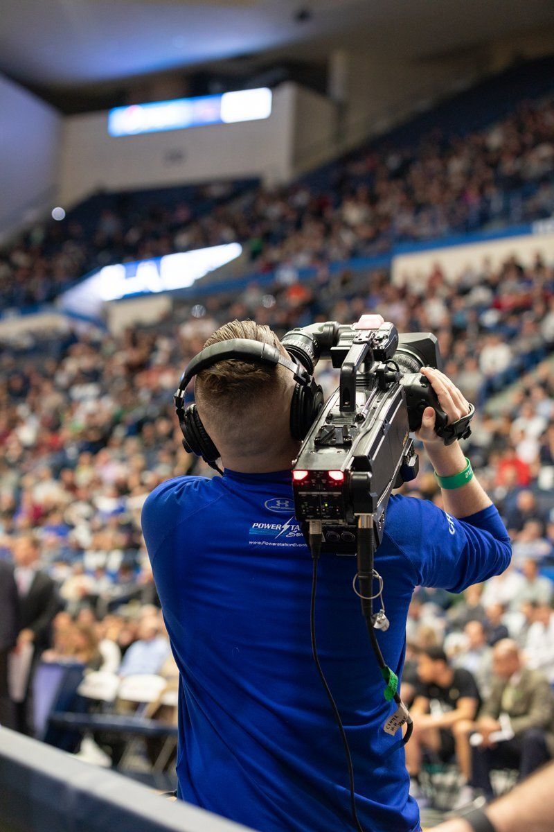 Camera operator filming a crowded indoor arena from behind, wearing a blue shirt and headset.