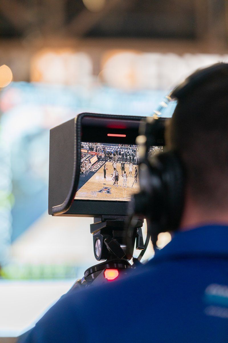 Camera operator filming a basketball game on a monitor, with a blurred viewer in the foreground