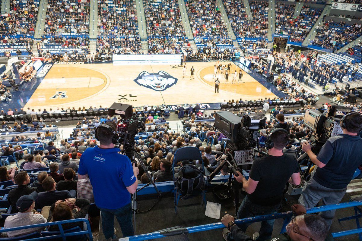 Wide view of a packed indoor basketball arena during a game, with fans and courtside cameras visible