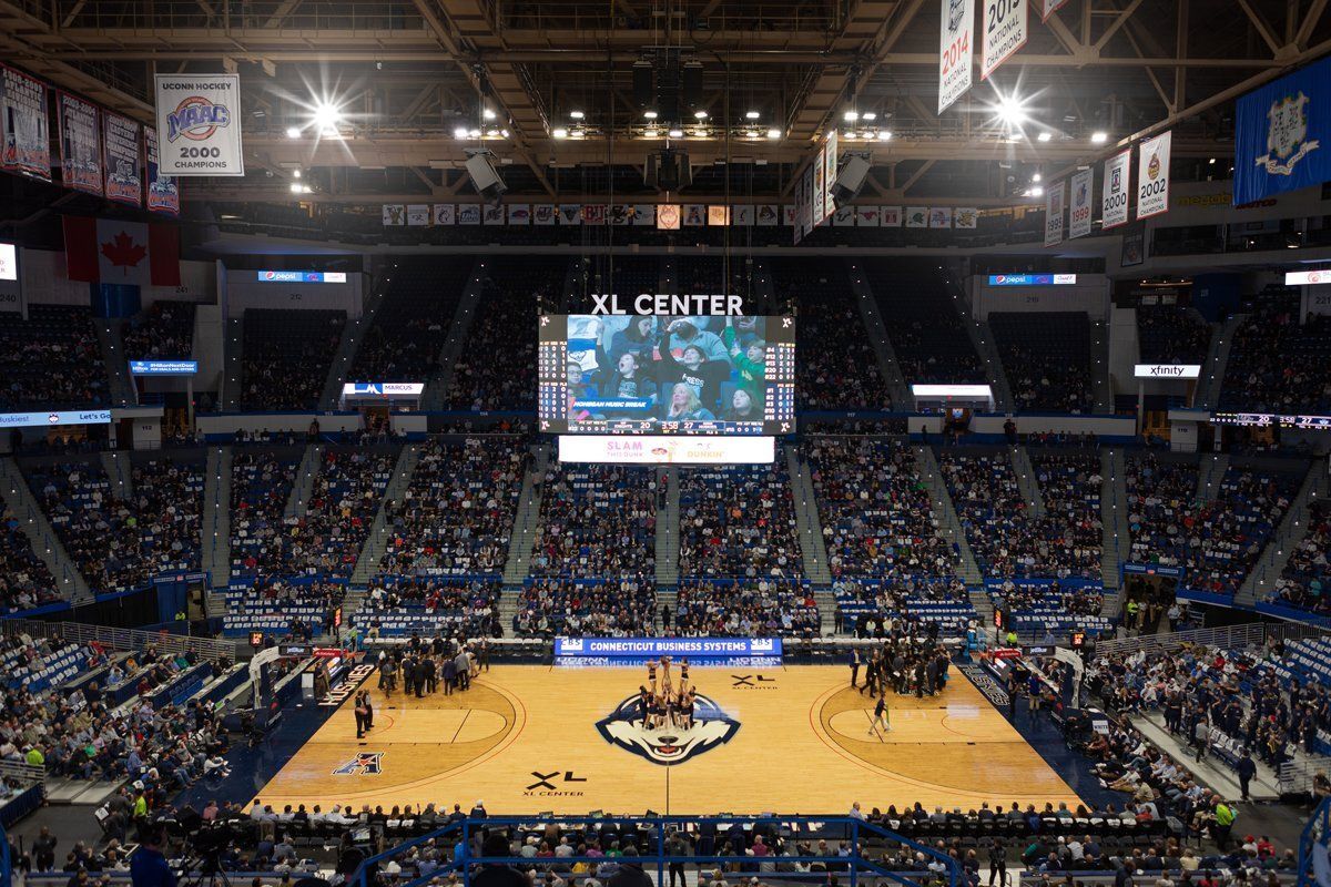Indoor basketball arena packed with fans, players on court, and a large scoreboard overhead.