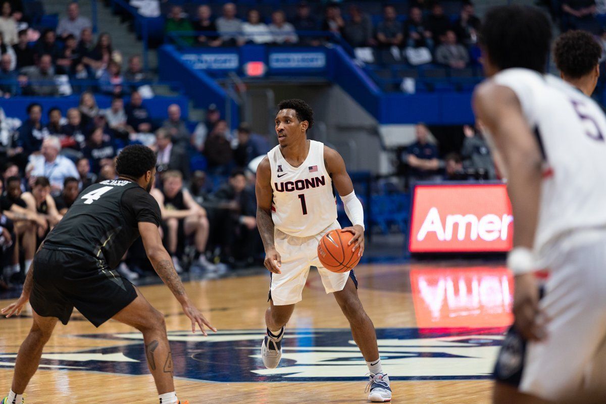 College basketball player dribbling on court while defender guards him, with UConn jersey visible in arena