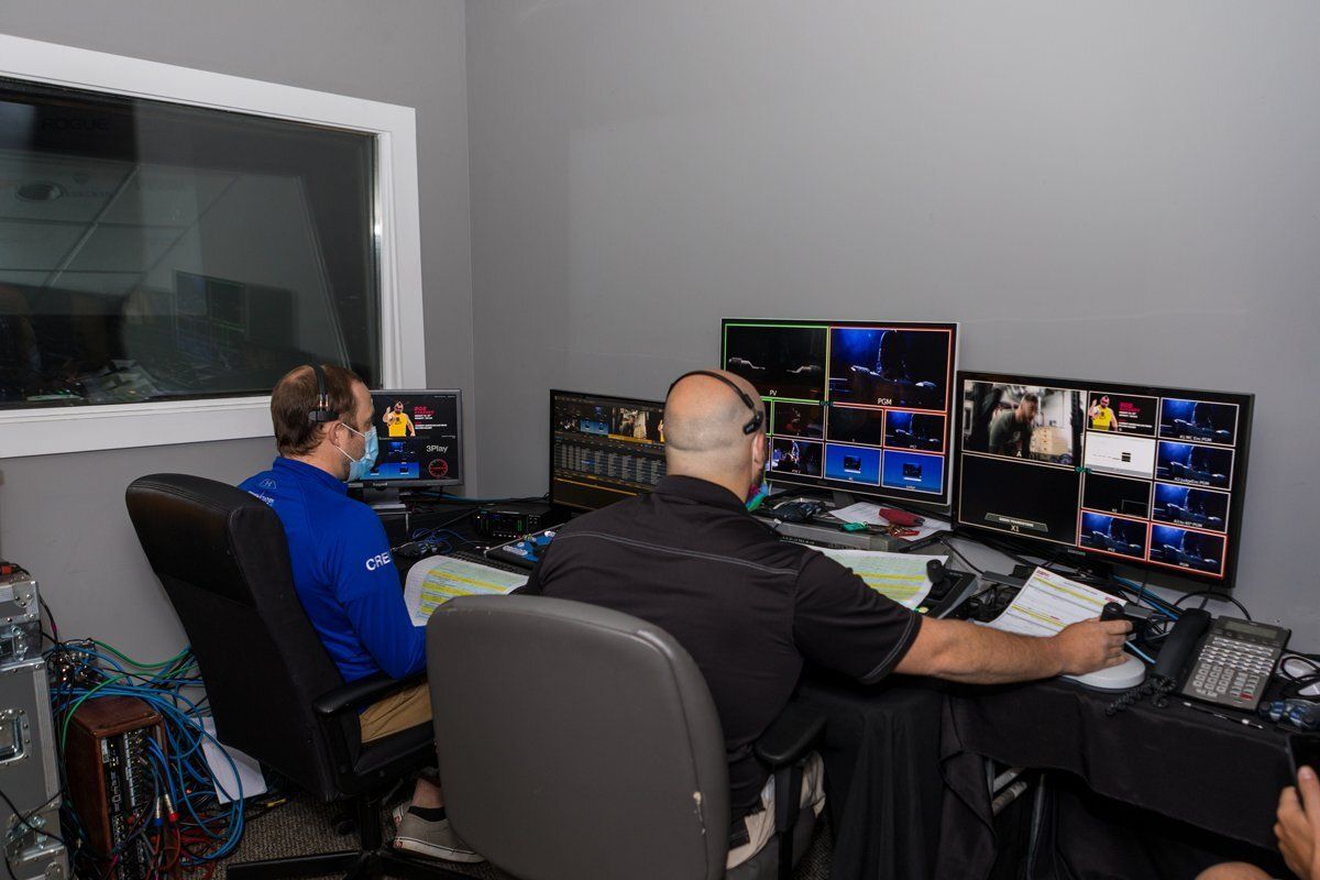 Two people operating multiple monitors in a control room, one in blue and one in black.