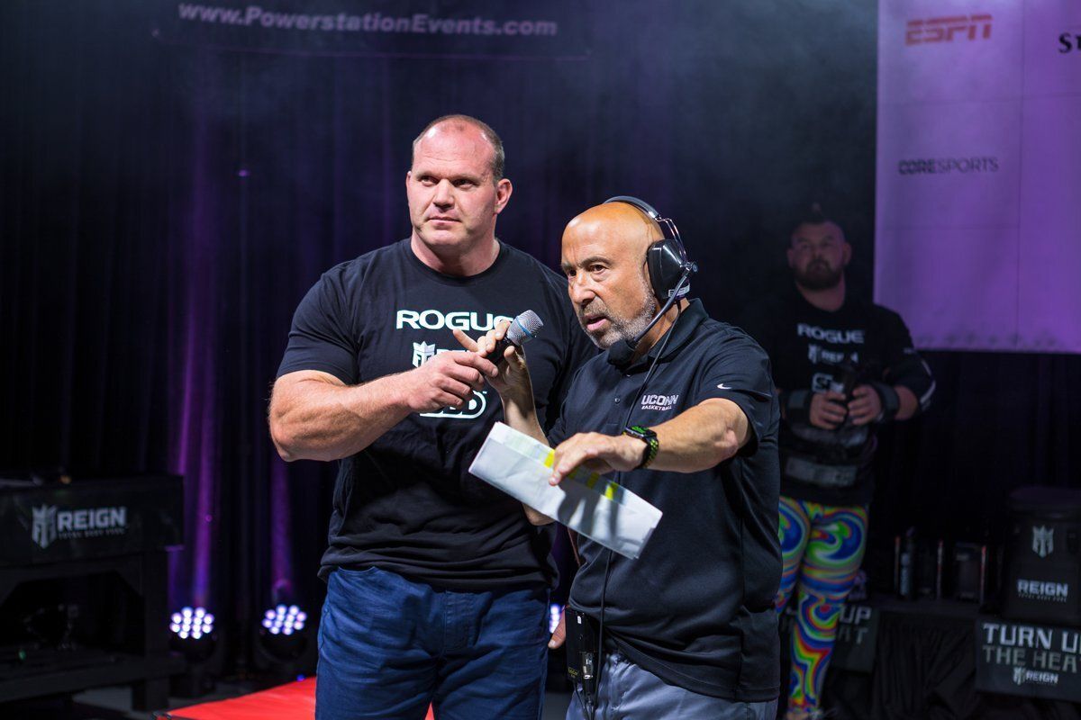 Two men in black shirts on a lit stage, one speaking into a microphone while the other stands beside him.