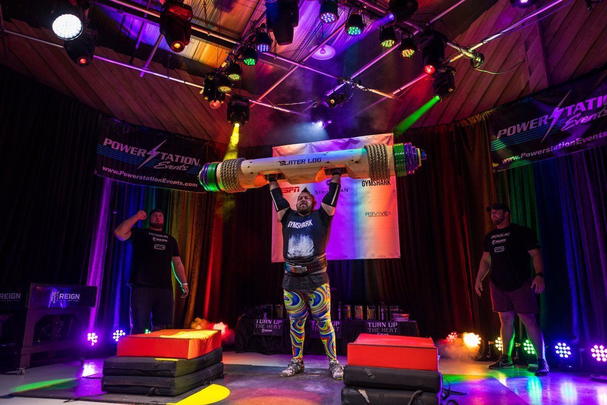 Performer lifting a large barbell overhead on a colorful lit stage with red and purple lights