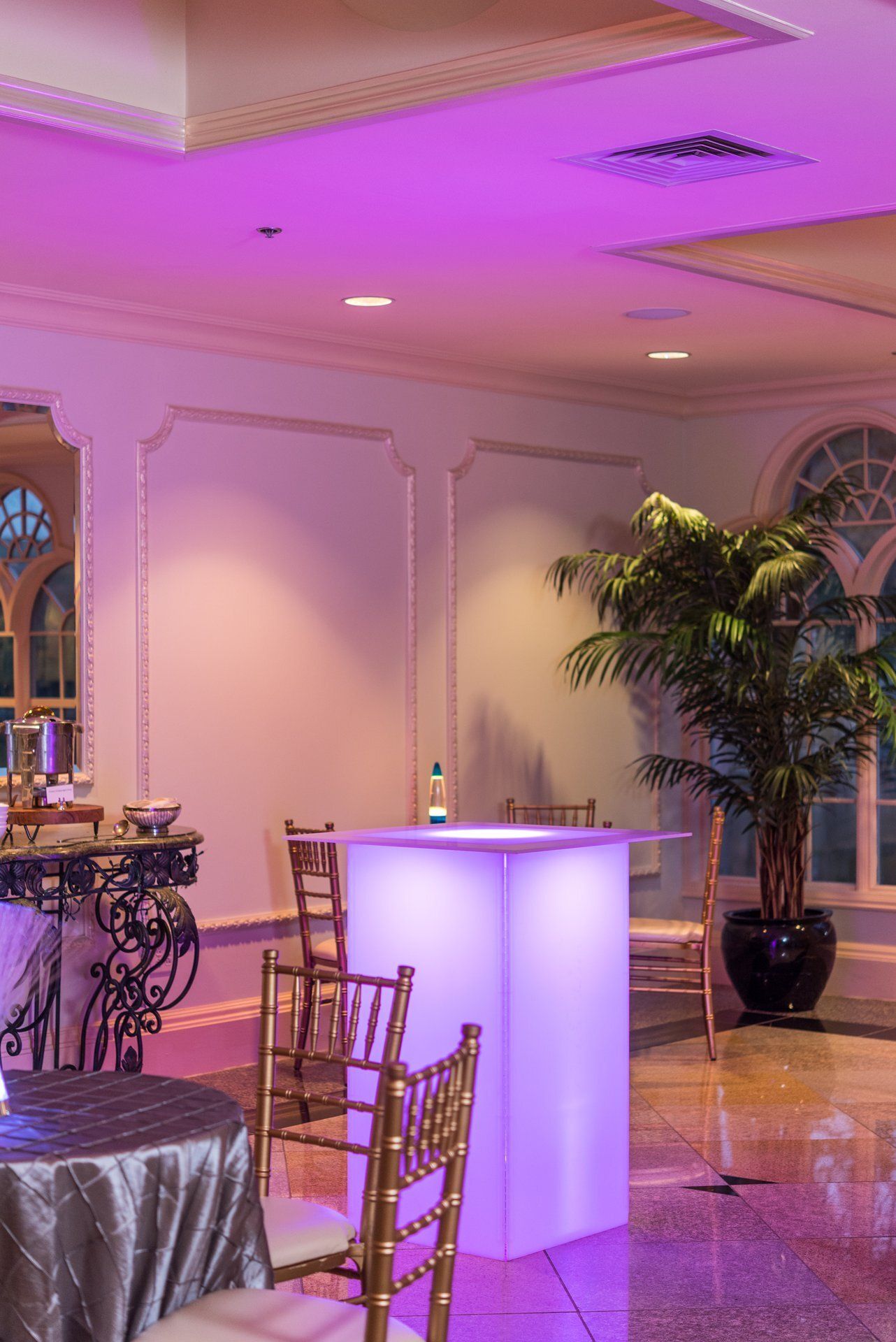 Elegant banquet hall with purple uplighting, white cocktail table, and gold chairs on a polished floor