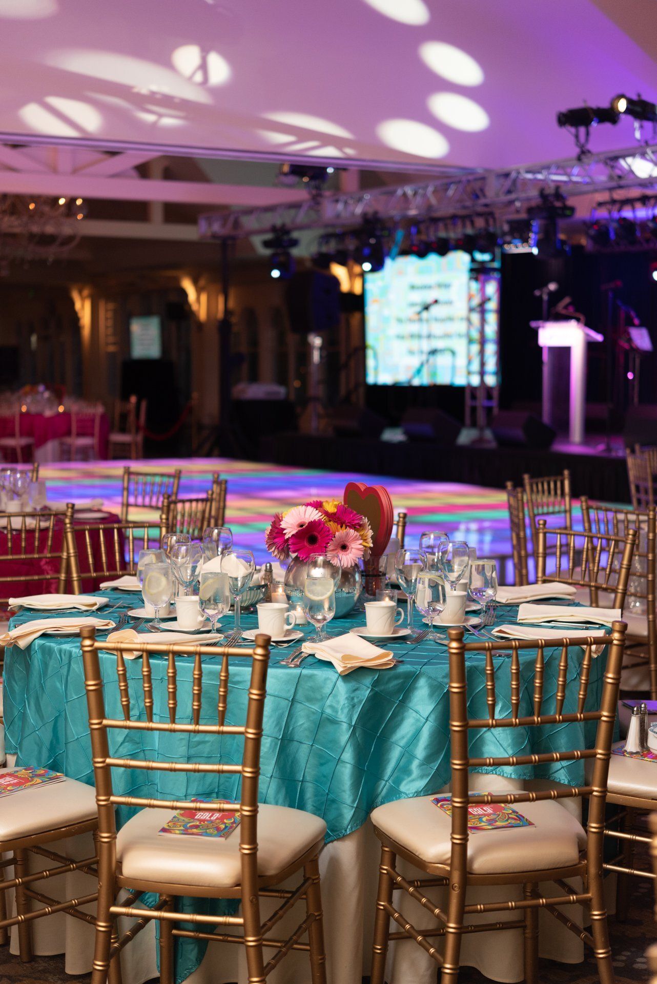 Elegant banquet hall with turquoise table settings, gold chairs, and a lit stage in the background