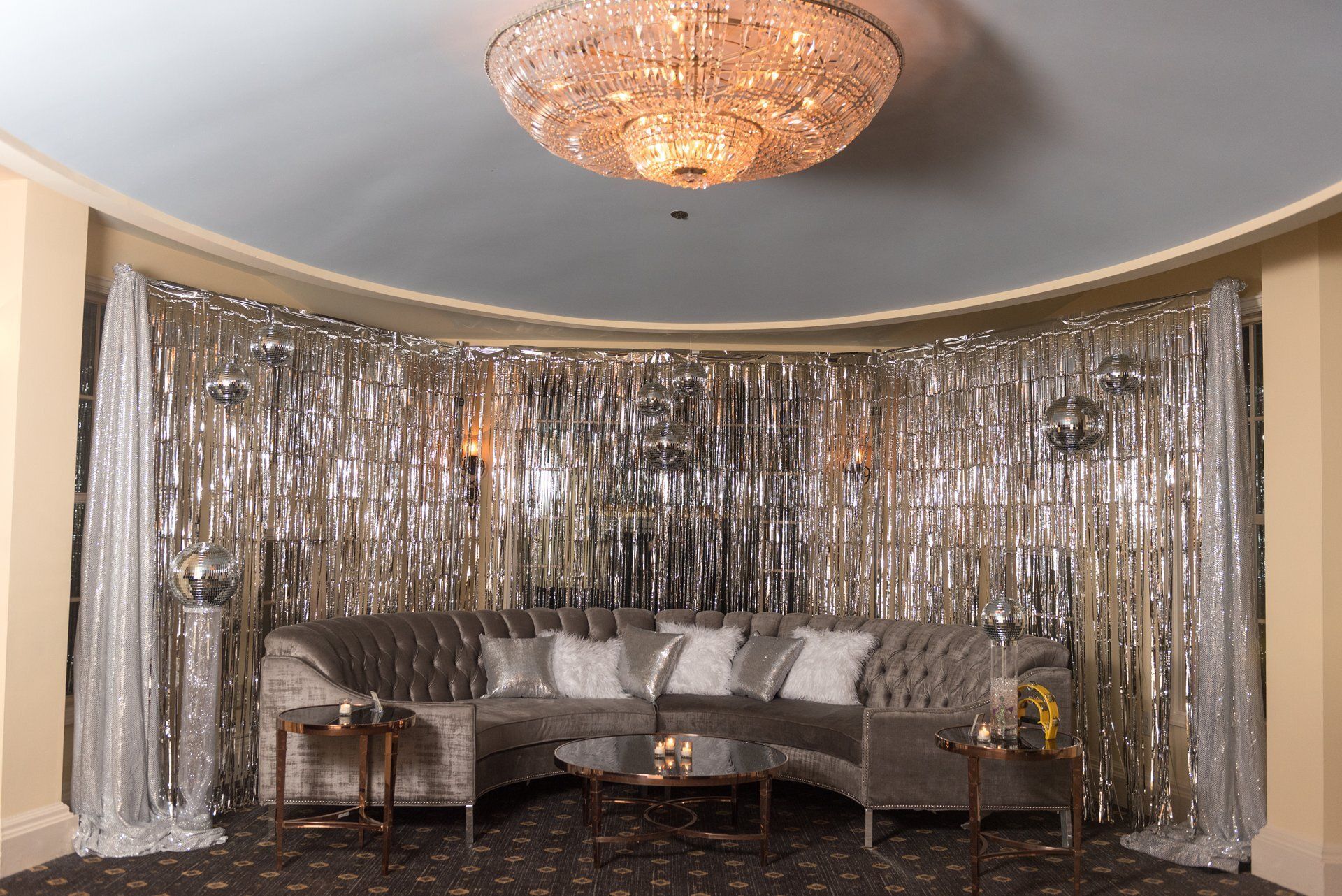 Elegant lounge with silver tinsel backdrop, curved sofa, side table, and chandelier under a blue ceiling.