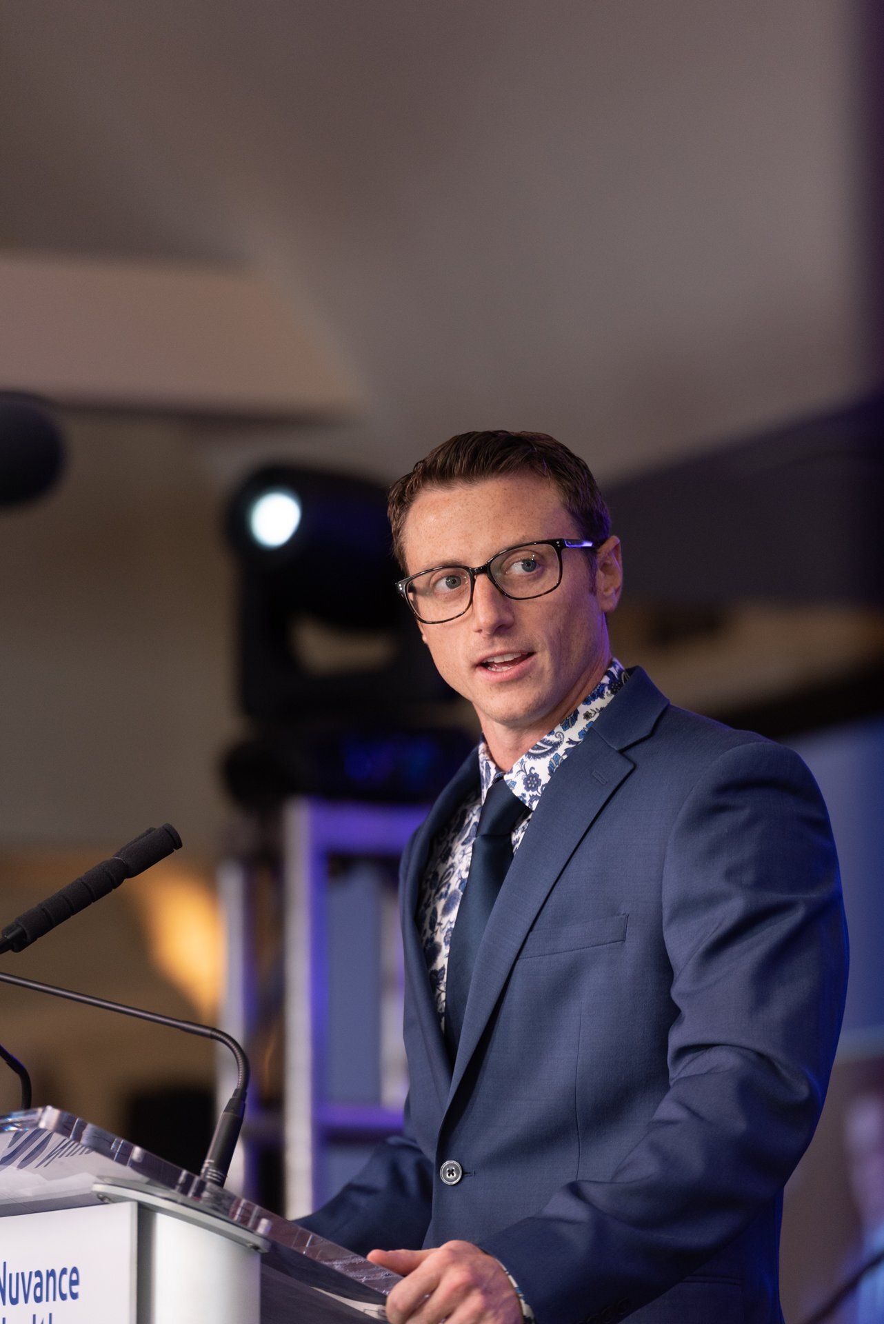 Man in a blue suit speaking at a podium under stage lights