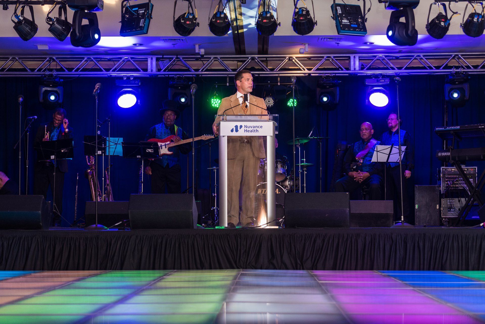 Speaker at podium on a lit stage with band equipment and colorful floor lights