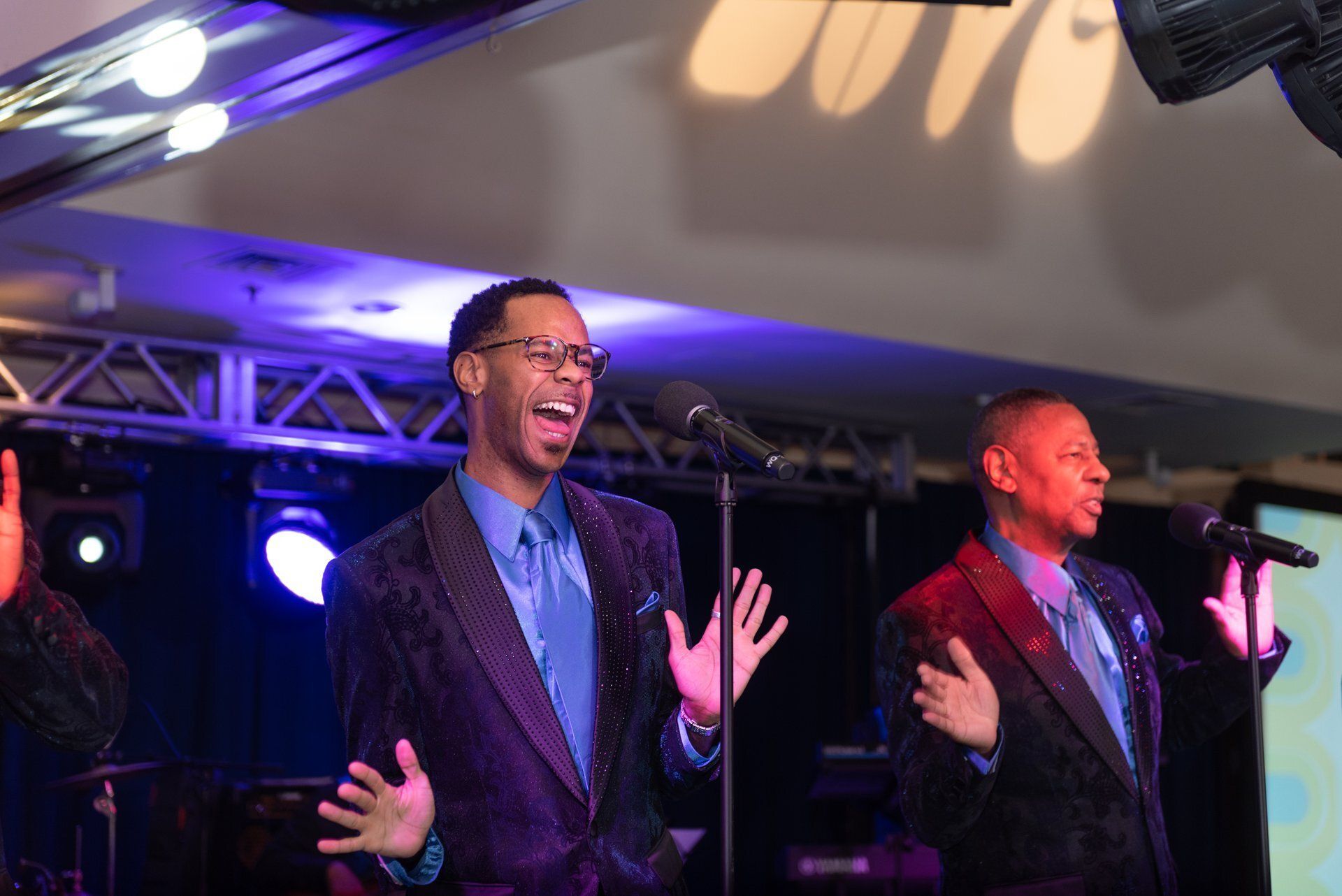 Two performers singing onstage under purple lights, one with hands raised in a lively pose