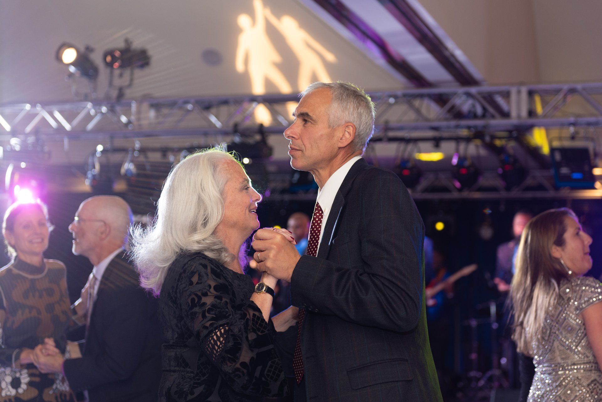 Couple dancing closely at a formal event, holding hands under purple stage lights.