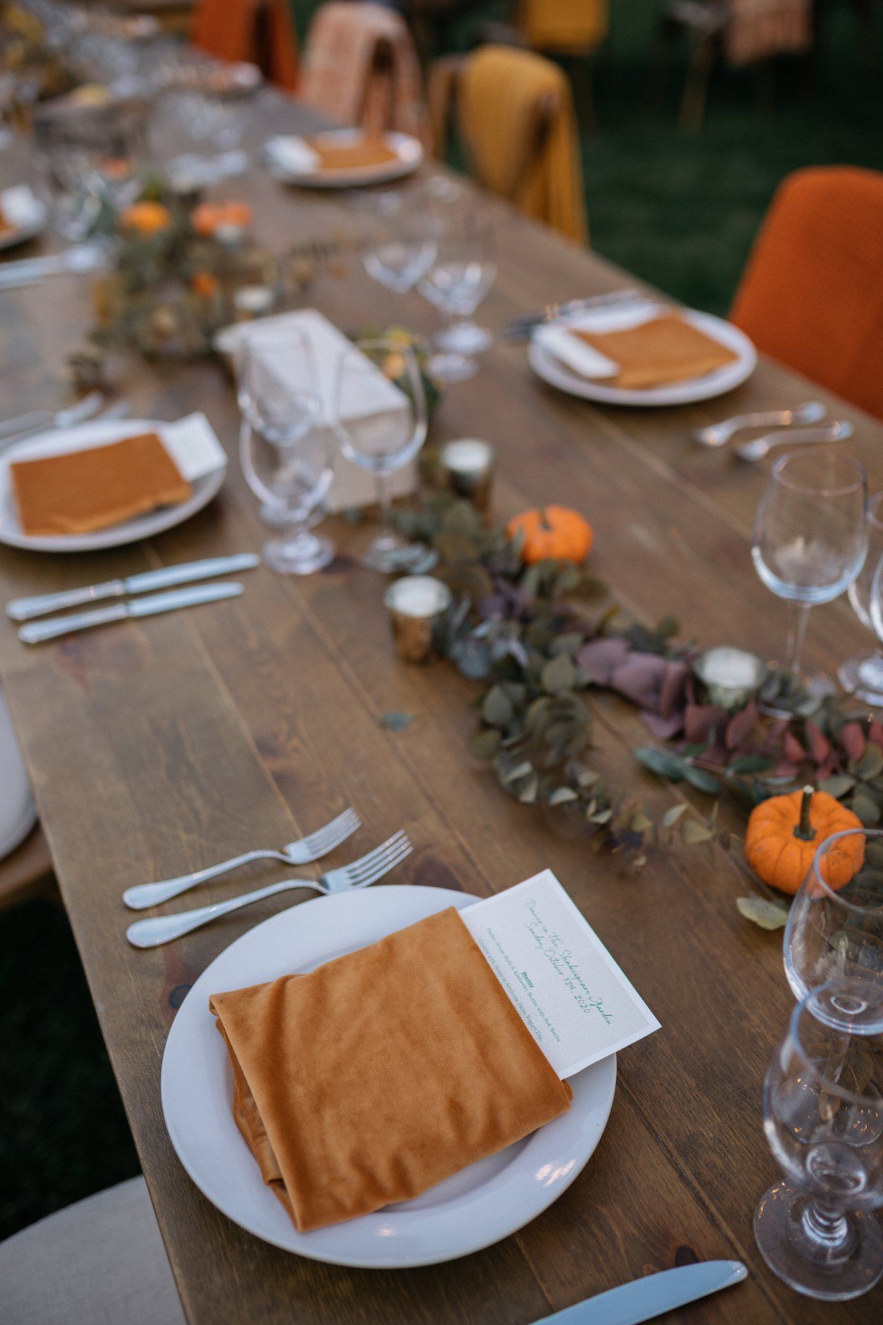 Long wooden dining table set with plates, glassware, napkins, and orange pumpkins as a fall centerpiece