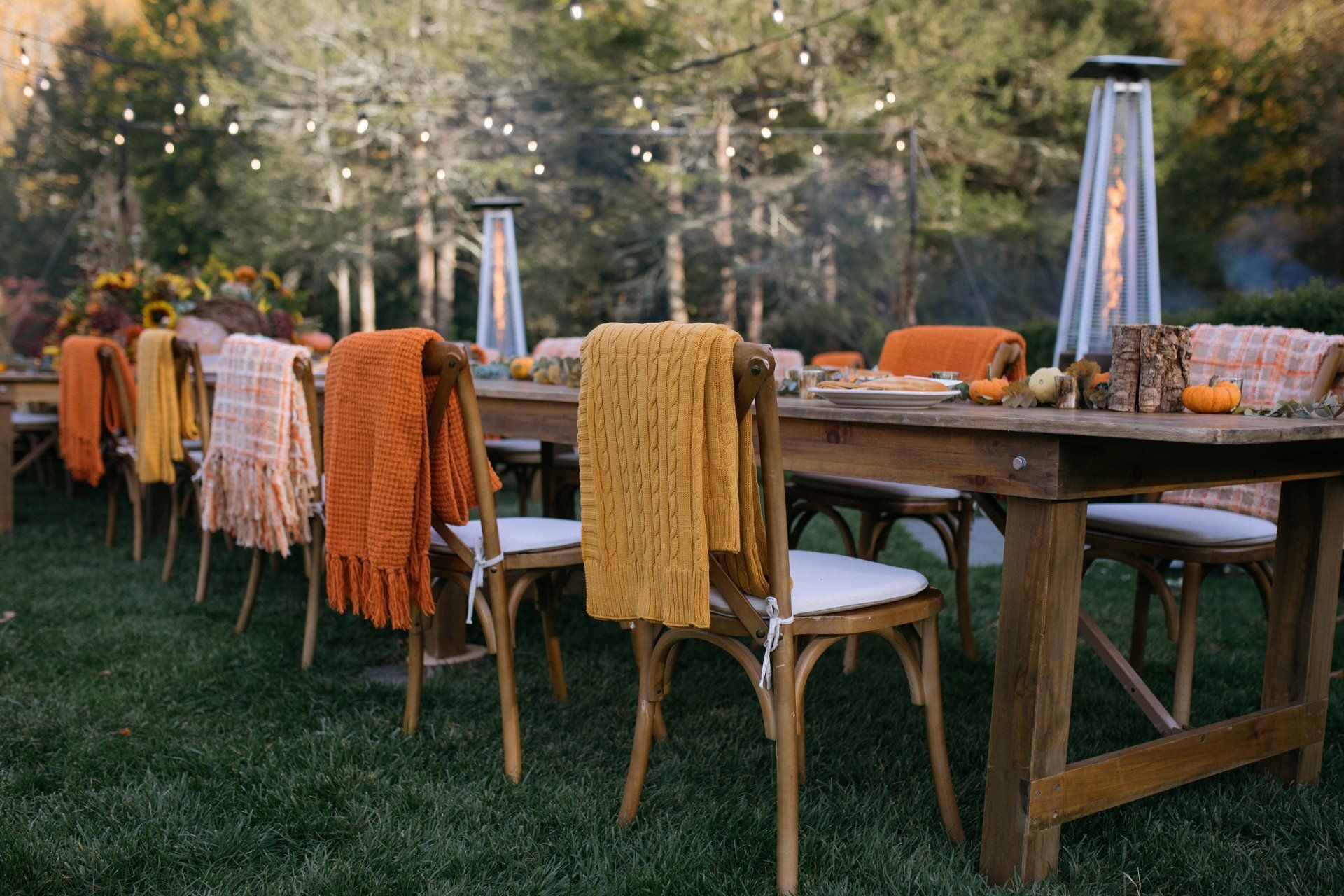 Outdoor dining table set with woven chairs draped in orange and yellow blankets on a grassy lawn.