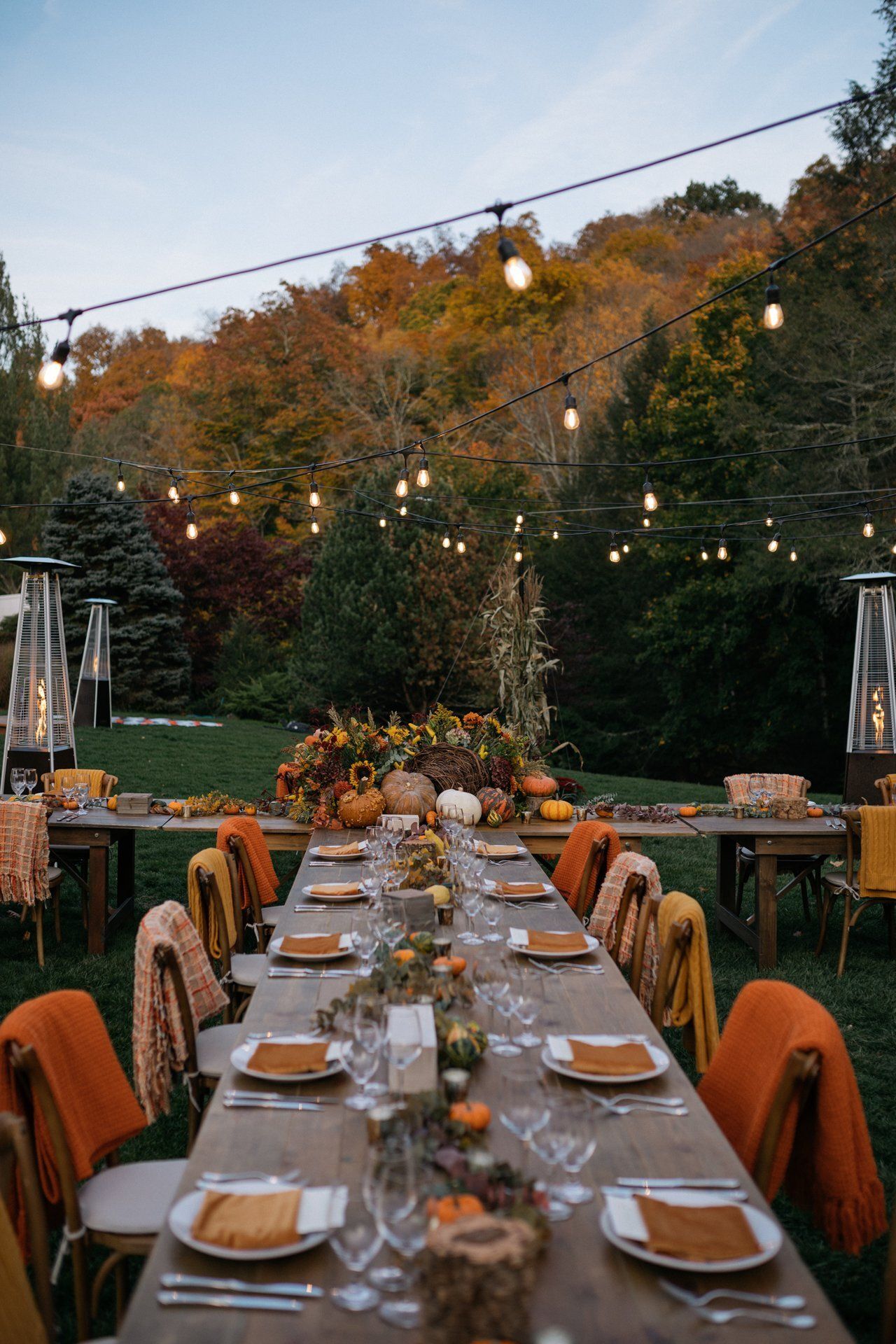 Outdoor dinner table set with candles and autumn foliage under string lights at dusk