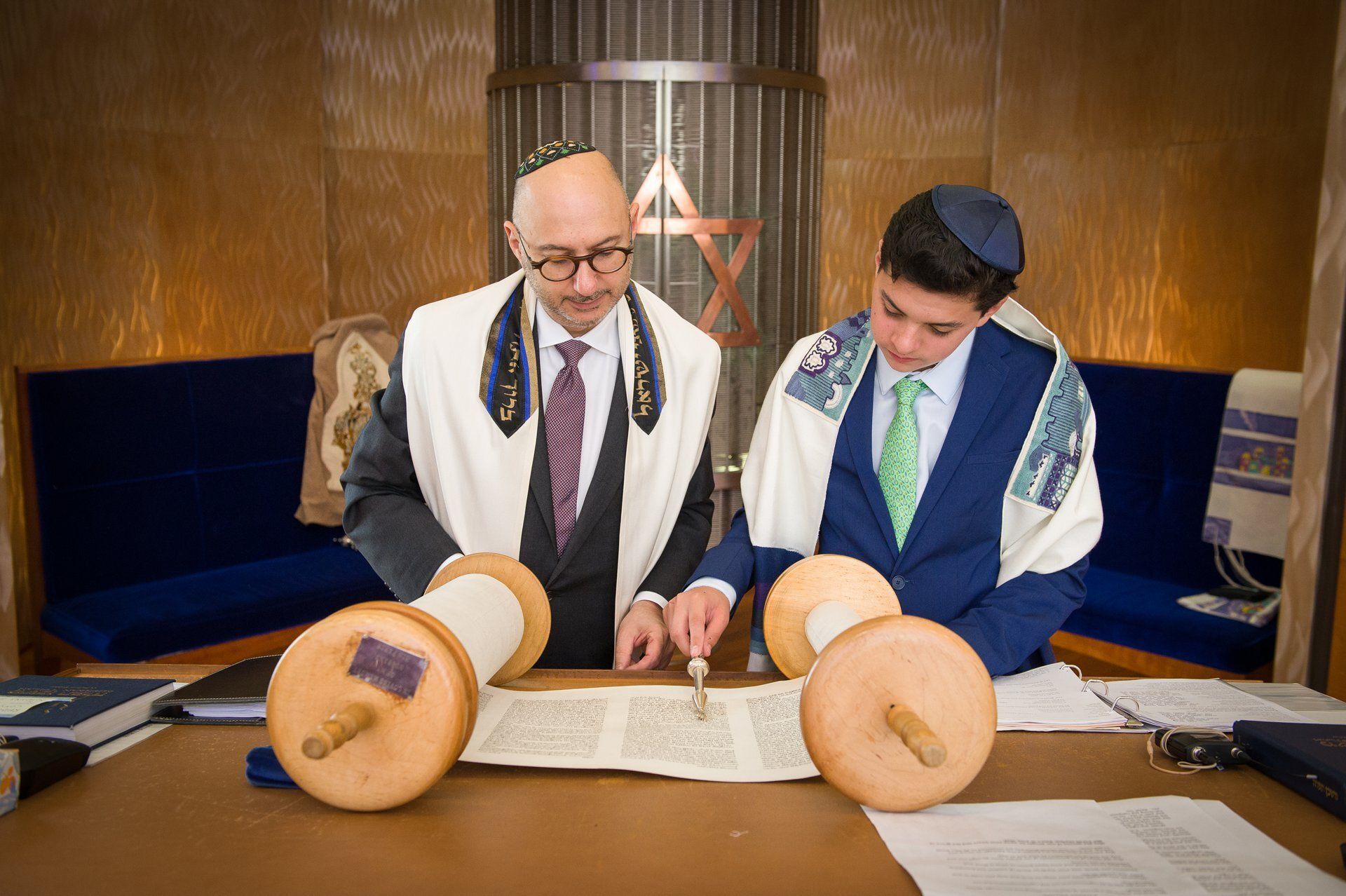 Two men in prayer shawls read from a Torah scroll in a synagogue.
