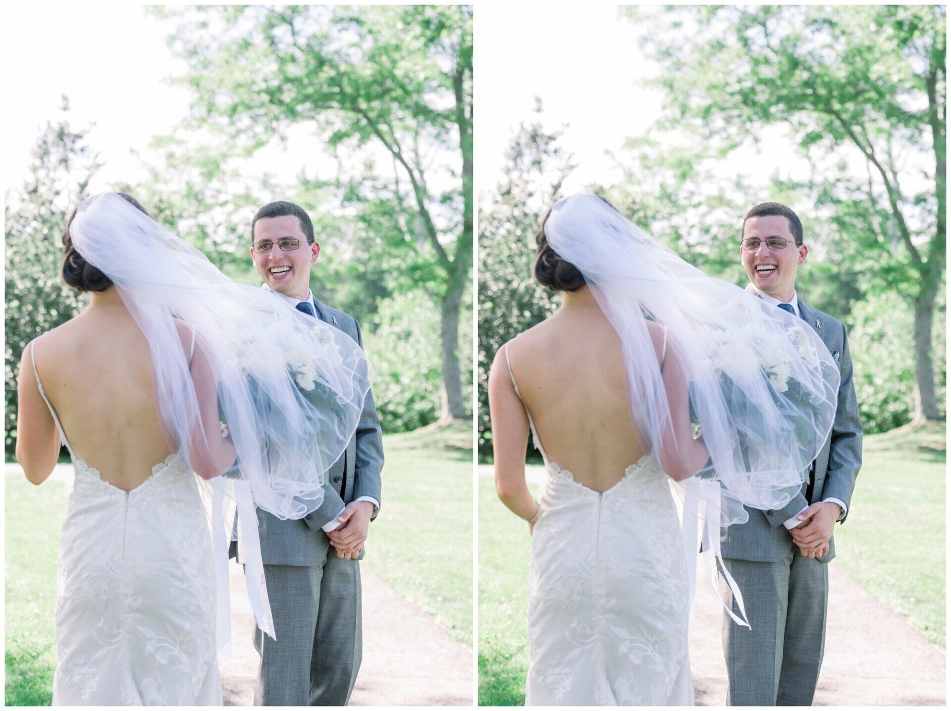 Bride in white dress and groom in gray suit smiling outdoors under trees, veil flowing in the wind